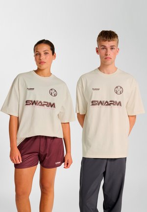 Beige oversized t-shirts med logo, kombineret med bordeaux shorts til venstre og grå træningsbukser til højre, hvilket fremhæver et sportigt design.