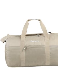 Beige sporttas met een gestructureerd hexagonaal patroon, dubbele handvaten, verstelbare schouderriem en een voorste ritszak. Logo weergegeven in wit.