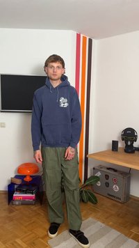 Marineblaue Kapuzenjacke mit weißem Grafikdesign, olivgrüne Cargo-Hose und schwarze Slipper, stehend in einem Raum mit bunten Akzentwänden.