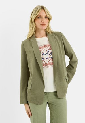 Femme blonde portant un blazer vert olive clair sur un t-shirt blanc imprimé et un pantalon olive assorti, debout devant un fond blanc uni.