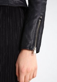 Rukáv černé kožené bundy s stříbrným zipem a detailním cvočkem. Tkanina se zdá být hladká s jemnou texturou.