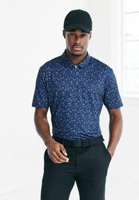 Marinblå polo med ett vitt abstrakt mönster, korta ärmar och krage med knappar, kombinerat med svarta byxor och en svart baseballkeps.