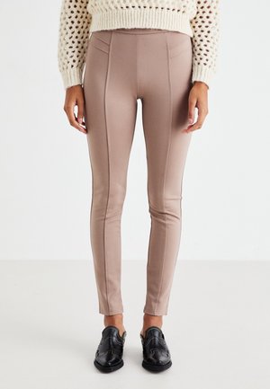 Leggings beiges ajustés avec coutures décoratives, texture lisse, taille haute, associés à des chaussures noires avec perforations décoratives.