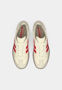 Ζευγάρι κρεμ αθλητικών παπουτσιών Adidas με κόκκινες ρίγες, δεμένα κορδόνια, φωτογραφία από πάνω σε λευκό φόντο.