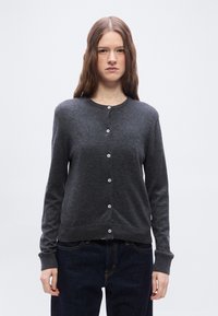 Νεαρή γυναίκα με μακριά κασταν ά μαλλιά που φοράει σκούρα γκρι ζακέτα με κουμπιά και σκούρο μπλε τζιν, στέκεται μπροστά σε ένα απλό λευκό φόντο.