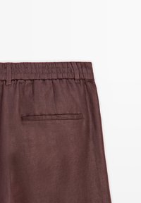Pantalon marron à taille élastique avec passants pour ceinture et une poche arrière passepoilée sur fond blanc.