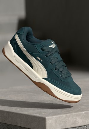 Baskets vert foncé en suède et mesh, avec des accents blancs et une semelle en caoutchouc marron. Les lacets et la languette sont rembourrés pour plus de confort.