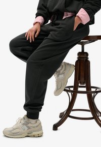 Sorte sweatpants med lommer og et logo, parret med lysebeige sneakers med grå accenter. En lyserød skjorte er lagd under.