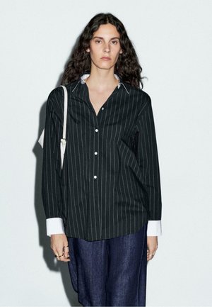 Vrouw met lang krullend haar draagt een zwart gestreept shirt met witte kraag en manchetten, donkerblauwe broek en een witte schoudertas.