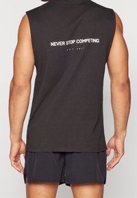 Chemise sans manches noire avec texte blanc "NEVER STOP COMPETING" et "EST 2017" au dos, en tissu léger.
