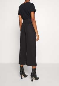Personne se tenant de dos, portant un pantalon large cropped noir, un haut noir à manches courtes et des bottines à talons hauts noires.