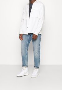 Bílá košile s knoflíky přes tmavě modrý henley, v kombinaci se světle modrými džínami a bílými teniskami. Dvě přední kapsy, volný střih.