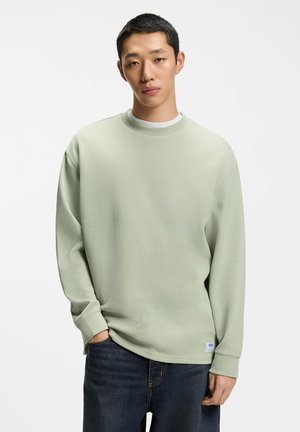 Joven con el pelo corto que lleva una camisa de manga larga texturizada de color verde claro y vaqueros azul oscuro, de pie con una mano en el bolsillo.