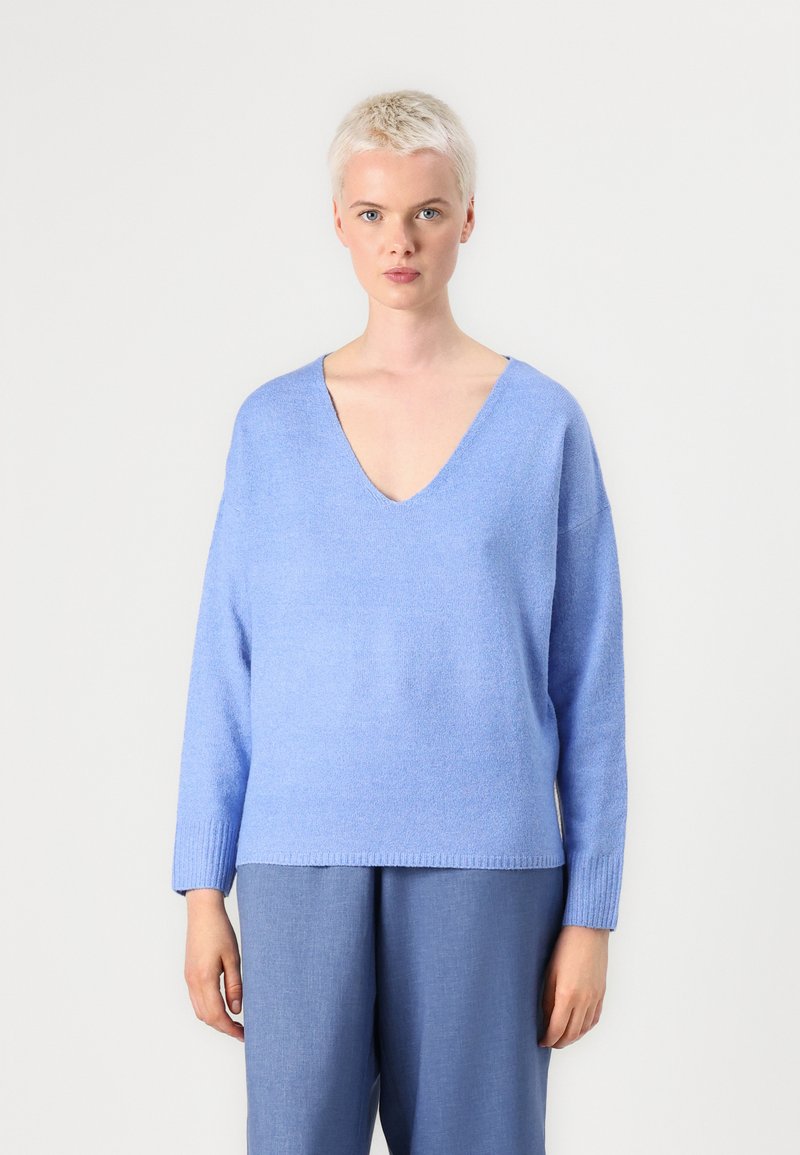 Pull en tricot bleu clair avec un décolleté en V profond, des épaules tombantes, des manches longues et un ourlet côtelé. Texture douce. Associé à un pantalon bleu assorti.