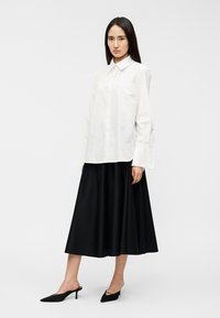 Λευκό πουκάμισο με κουμπιά και μακριά μανίκια, συνδυασμένο με μαύρη πλισέ μίντι φούστα. Φορεμένο με μαύρα, μυτερά παπούτσια με τακούνι.