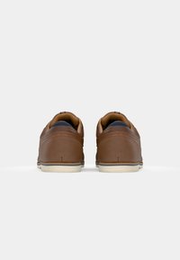 Chaussures en cuir marron avec une texture lisse, présentant une forme arrondie et des semelles blanches contrastées. Le design inclut des détails de couture subtils.