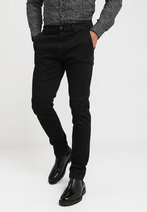 Man draagt strakke zwarte broek, gepoetste zwarte laarzen en een zwart-wit polkadot shirt met lange mouwen, handen in de zakken.