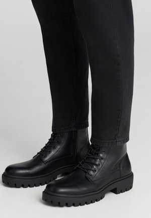 Stivaletti alla caviglia in pelle nera con lacci, cerniera laterale e suola spessa scanalata, indossati con pantaloni neri.