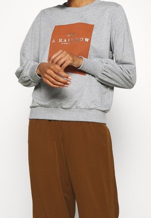 Grå sweatshirt med en rostorange rektangulär grafik som står "VAR EN REGNBÅGE" och elastiska ärmslut, tillsammans med bruna byxor.