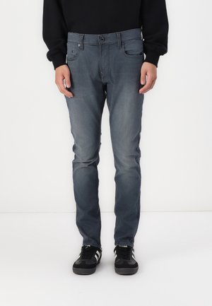 Persoon draagt vervaagde blauw slim-fit jeans en zwarte sneakers met witte strepen, staand tegen een effen witte achtergrond.