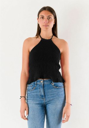 Donna con lunghi capelli castani indossa un top nero a fascia e jeans blu, in piedi davanti a uno sfondo bianco uniforme, rivolta in avanti con espressione neutra.