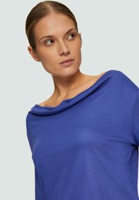 Blauwe off-shoulder top van zachte stof, met een gedrapeerde halslijn en een losse pasvorm, matte textuur zonder zichtbare patronen.