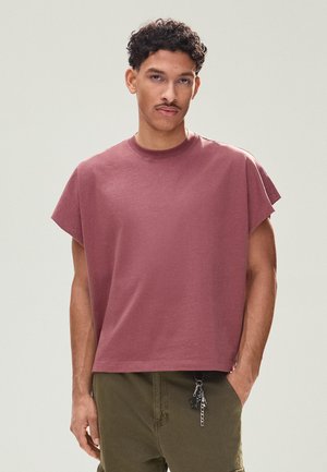 Mladý muž s krátkymi kučeravými vlasmi, oblečený v voľnom fialovom tričku s krátkym rukávom a olivovozelených nohaviciach s kľúčenkou visieťou z vrecka.