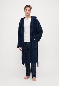 Marineblå, myk badekåpe med hette, åpen front og to lommer, brukt over en hvit henley-skjorte og rutete pysjbukser i marineblått og hvitt.