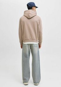 Sudadera beige con capucha y cordones, combinada con unos jeans de pierna ancha azul claro que muestran una textura desvaída y costuras. Una gorra azul completa el look.