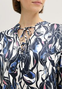 Chemise avec un col noué, présentant un motif abstrait bleu, blanc et rose sur un fond clair. Fabriquée en tissu léger.