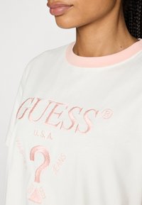 Tricou alb din bumbac cu un guler roz ribat, având textul brodat proeminent în roz "GUESS" și elemente suplimentare de branding în roz deschis.