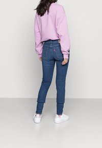 Mujer de espaldas vistiendo un sudadera ligera morada, jeans de cintura alta azul y zapatillas blancas, sobre un fondo claro y liso.