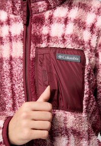 Mano che apre una piccola tasca con cerniera su una giacca in pile Columbia a scacchi bordeaux e rosa.