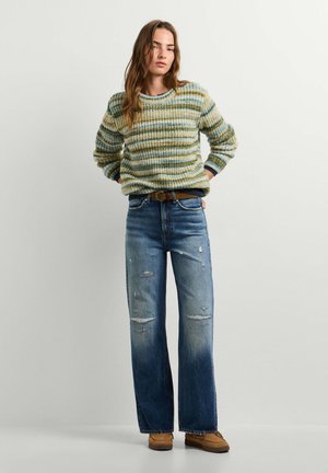 Femme portant un pull tricoté rayé, un jean large usé, une ceinture marron et des chaussures couleur fauve, debout devant un fond clair uni.