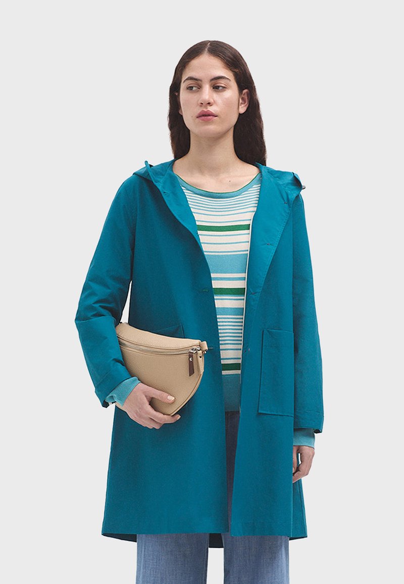 Impermeabile verde acqua con cappuccio e chiusura a bottone, dotato di due tasche frontali, indossato sopra una maglietta a righe, abbinato a una borsa a tracolla curvata beige.