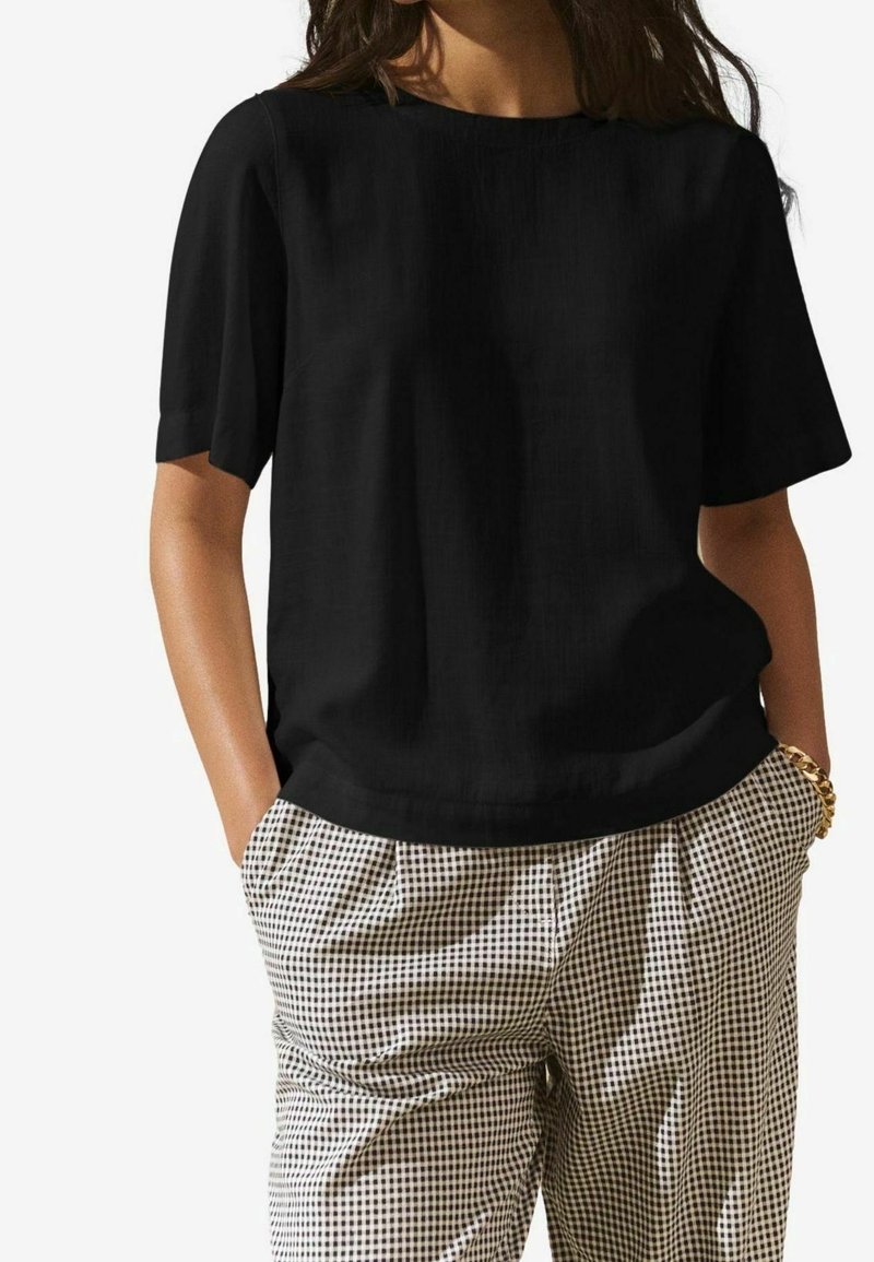 Femme portant un haut noir à manches courtes et un pantalon à carreaux beige et noir taille haute, les mains dans les poches.