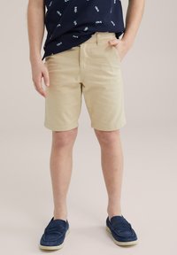 Kaki shorts med en slät textur, rak framkant och uppvikta fållar. Kombinerat med en marinblå skjorta med ett ljust mönster. Blå mockaskor.
