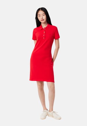 Junge Frau steht mit den Händen hinter dem Rücken, trägt ein rotes Polokleid und weiße Turnschuhe, vor einem einfarbigen hellen Hintergrund.