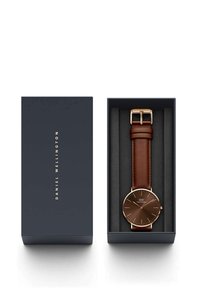 Brown leather strap watch in a blue box. Round brown dial with gold markings and hands. Closed buckle with gold hardware. Soft interior.