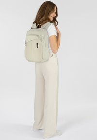 Zaino beige chiaro con forma arrotondata, realizzato in tessuto testurizzato, dotato di una tasca anteriore con zip e compartimenti laterali; logo del marchio visibile.