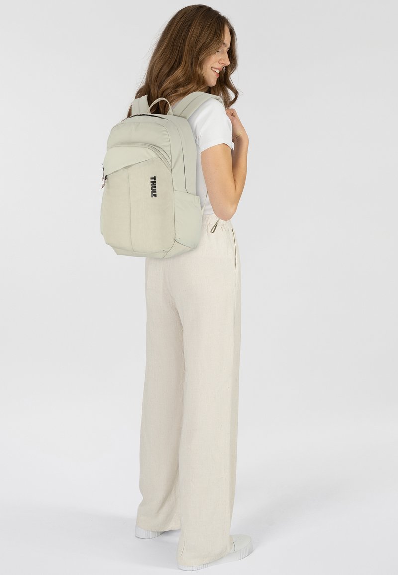 Zaino beige chiaro con forma arrotondata, realizzato in tessuto testurizzato, dotato di una tasca anteriore con zip e compartimenti laterali; logo del marchio visibile.