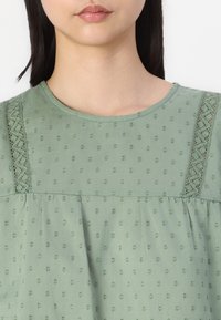 Blusa verde pallido con pois testurizzati, dettagli in pizzo sulle spalle, scollatura rotonda e vestibilità ampia attraverso il corpo.