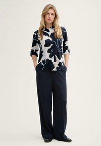 Overhemd met knopen in zwart-wit abstract patroon met blauwe accenten; gecombineerd met marineblauwe broek met wijde pijpen; stof lijkt glad en lichtgewicht.