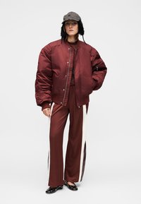 Veste bomber bordeaux à silhouette oversize, associée à un pantalon à jambes larges assorti avec des bandes latérales crème et brun foncé. Casquette grise.