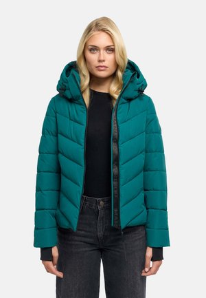 Femme aux longs cheveux blonds portant une doudoune turquoise avec capuche, un t-shirt noir et un jean foncé taille haute, regardant devant avec une expression neutre.