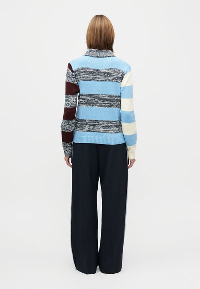 Strikket genser med flerfargede striper i blått, grått, burgunder og krem. Har høy krage og løs passform, kombinert med vide svarte bukser.