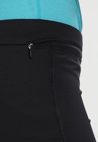 Černé legíny se širokým pasem, s malou zipovou kapsou a hladkým, elastickým materiálem. Nad nimi je vidět světle modrý top.