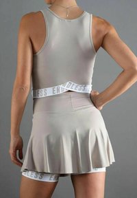 Mujer vestida con una blusa sin mangas beige y una falda a juego, con bandas elásticas blancas etiquetadas como "ENDLESS," de pie con una mano en la cadera.