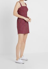 Vestito a quadri rosso e nero con scollatura quadrata, spalline sottili e chiusura frontale con bottoni, abbinato a sneakers bianche. Tessuto liscio.