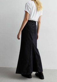 Jupe maxi en denim noir avec une forme évasée et une fente arrière, comportant deux poches arrière et des coutures contrastées. Portée avec un t-shirt blanc.